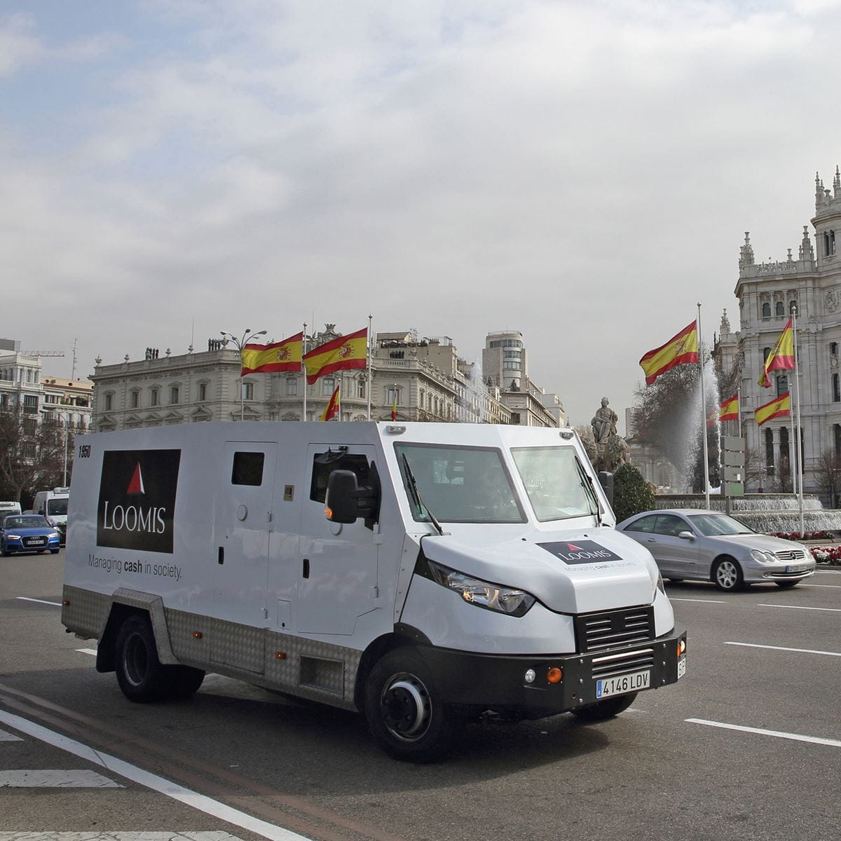 Carretera con banderas de España al fondo y un coche blindado de Loomis Pay, visible de frente a trasera con los logos en ambos lados.